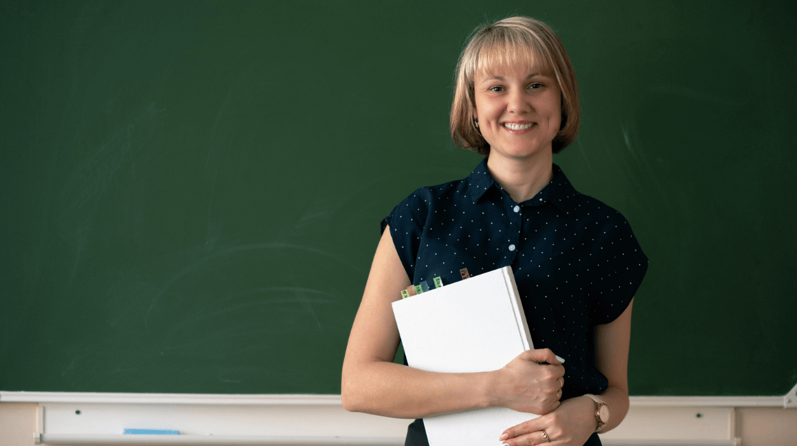 Uśmiechnięta nauczycielka stojąca przed tablicą, trzymająca w rękach segregator z dokumentami